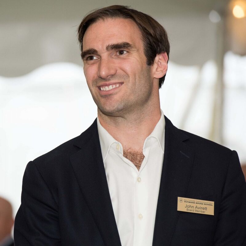 The image shows a man with short brown hair, wearing a white shirt and a dark blazer. He has a name tag that reads "John Avirett". He is smiling and looking slightly to the side. The background is blurred, suggesting an indoor event.